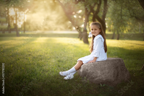 a beautiful little girl in a white dress is sitting on a rock in a sunlit park