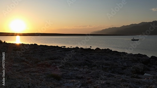 Evening landscape with setting sun, rocky beach silhouette in forefront, bay with single fisherman boat and Velebit mountain in background. Clear skies with some clouds. Location near Razanac, Croatia