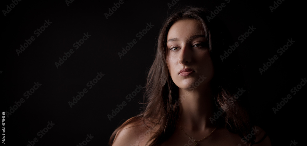 Studio portrait of a young Caucasian woman with long brown hair, looking serious with bare shoulders against a dark background Studio portrait of a young Caucasian woman with long brown hair, looking serious with bare shoulders against a dark background