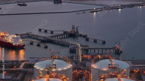 LNG tanker docked at an offshore terminal with multiple pipelines, storage tanks, and an industrial complex. Captures global energy logistics