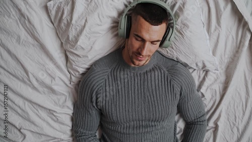 Handsome man relaxing on bed with headphones wearing sweater peaceful music moment