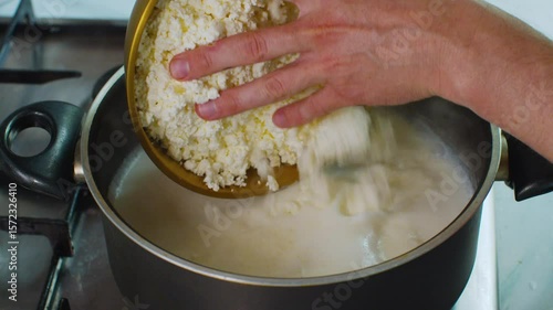 Pouring curd into a pot of boiling milk for homemade cheese preparation. Close-up of traditional dairy processing in a home kitchen
