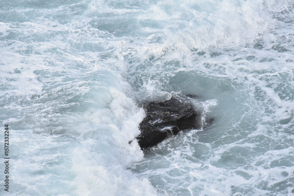 Fototapeta premium Powerful waves crash over a dark rock beneath the swirling turquoise waters of Nazaré, Portugal — nature’s raw energy frozen in motion.