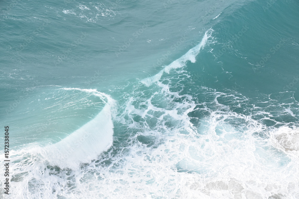 Fototapeta premium A powerful turquoise wave crashes onto the shore, captured in stunning detail from above. The seafoam swirls in vibrant white against the crystal-clear blue water