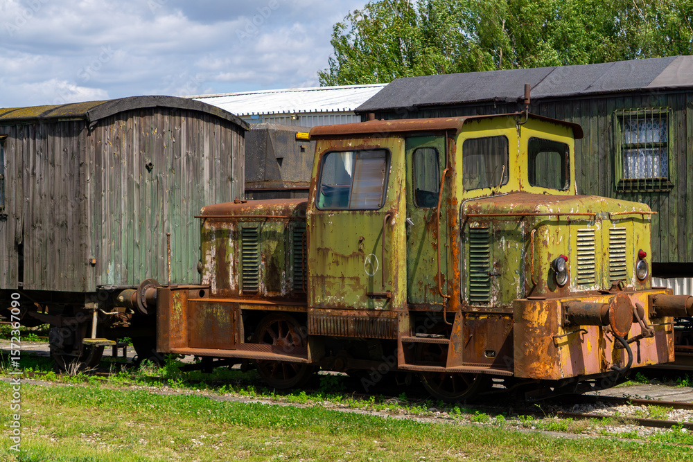 Obraz premium Rusty narrow-gauge locomotive next to old wooden railcars on a side track