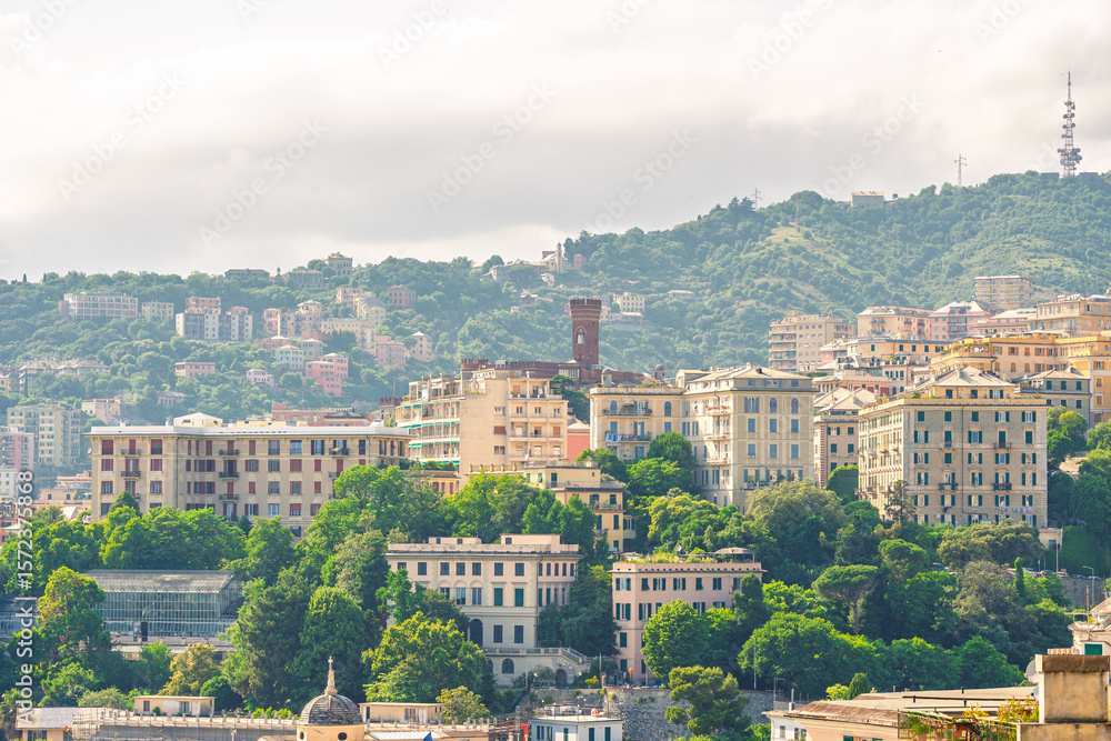 Fototapeta premium View over Genoa’s hillside neighborhoods with layered residential buildings, a red tower, and lush greenery under soft summer light