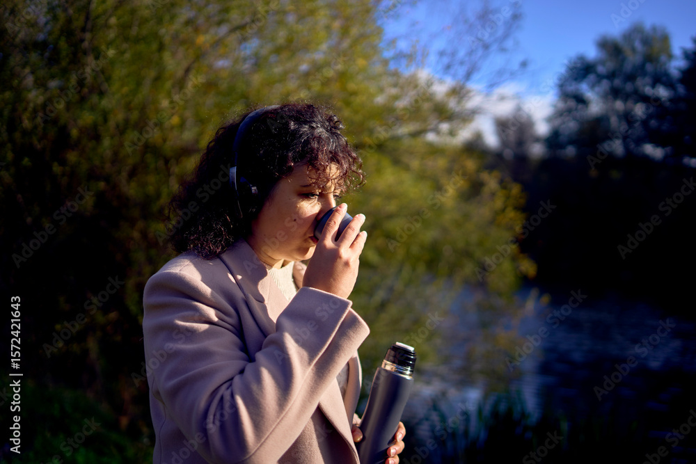 Obraz premium middle-aged woman in wireless headphones drinking coffee, tea from a thermos on a walk in the autumn forest