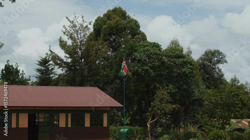 New single-storey building in savannah settlement with country's flag on flagpole and water tank at entrance. Stunning diversity of vegetation against a cloudy sky