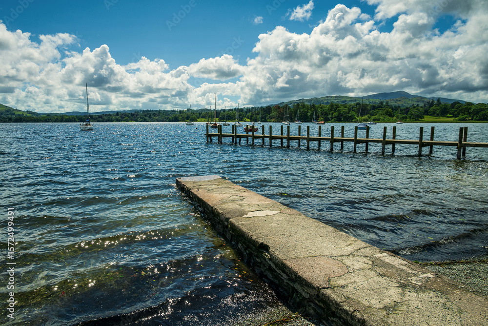 Fototapeta premium Wind-driven waves glisten in the summer light on Windermere’s rippling surface, seen from a concrete slipway at the shore, capturing the lively energy of a breezy holiday day in the Lake District.