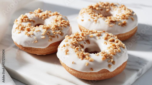 Three delicious donuts with white glaze and crushed nuts topping, arranged on a white marble surface in bright natural light, evoking sweetness and indulgence.