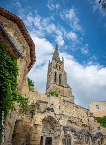A striking view of the Saint-Emilion troglodytic church towers over ancient stone structures, embraced by green vines, under a vibrant spring sky in the Bordeaux wine region.