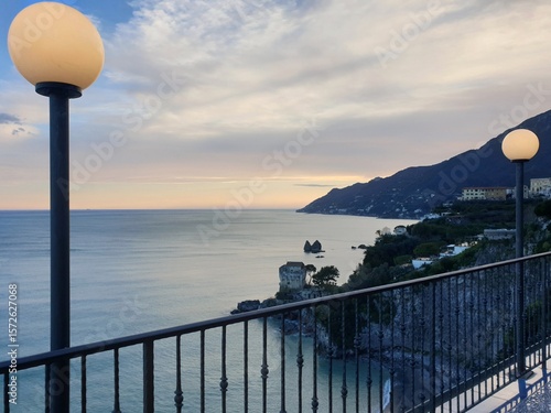 Old fashioned lamps with round globes are part of a verandah which overlooks hills surrounding the Gulf of Naples. Below is an old fortification. The sea is calm and the clouds show the colours of sun