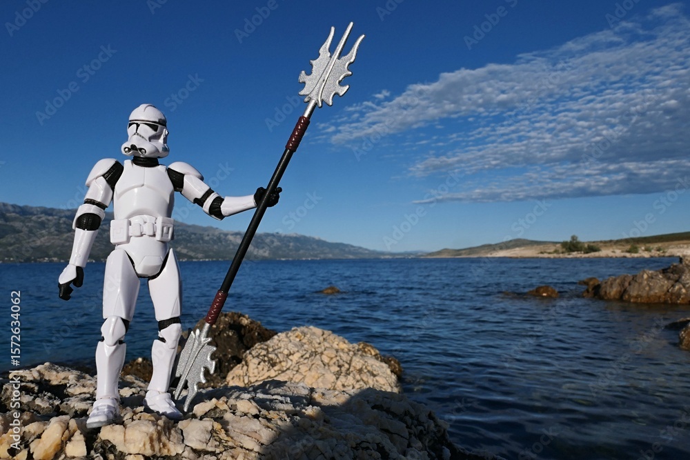 Naklejka premium Star Wars action figure of Clone trooper in white armor and helmet, holding spiked long asian staff in position like Poseidon with triton. Croatian rocks on bay shore, blue skies with clouds.
