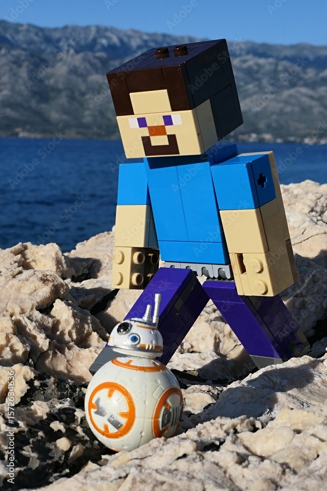 Naklejka premium Closeup view of LEGO Minecraft figure of Steve on walk with small model os Star Wars astromech droid BB-8 on rocky Croatian beach. Bay water, Velebit mountain and blue skies in background.
