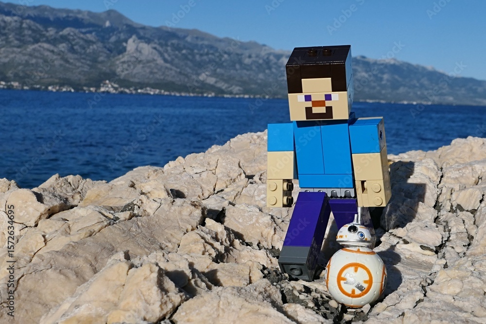 Naklejka premium LEGO Minecraft figure of Steve on walk with small model os Star Wars astromech droid BB-8 on rocky Croatian beach. Bay water, Velebit mountain and blue skies in background.