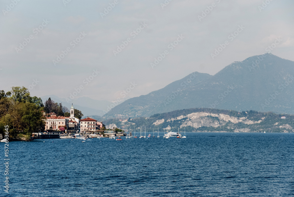 Naklejka premium Scenic Lake Maggiore vista featuring sailboats gliding over azure waters, Belgirate nestled amid dramatic mountain peaks, capturing classic Italian lakeside tranquility