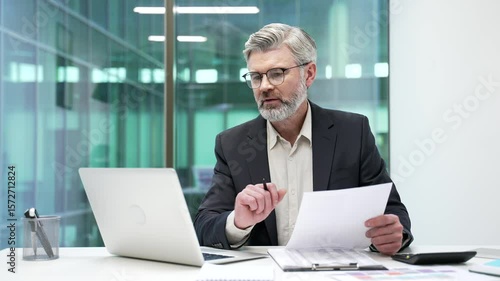 Joyful mature businessman is satisfied with results of a financial report looking at documents and computer screen sitting in business office. Manager in formal suit is happy with positive indicators