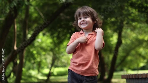 A young child with medium-long hair, wearing an orange polo shirt, stands in a green park or forest