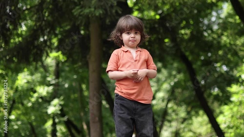 A young child with medium-long hair, wearing an orange polo shirt, stands in a green park or forest. The child gestures with curiosity and a playful expression. 