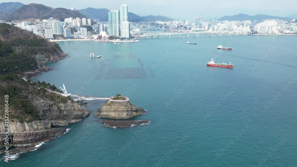 Drone aerial view in South Korea countryside flying over the clear blue sea of Busan next to a green mountain, ships and city in the background sunny day Drone aerial view in South Korea countryside flying over the clear blue sea of Busan next to a green mountain, ships and city in the background sunny day