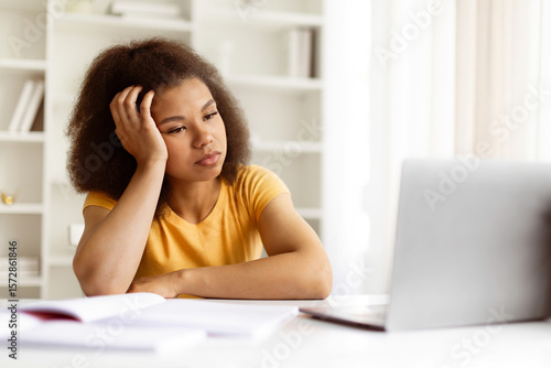 Bored young black woman resting her head on hand while staring at laptop. Tired of screen time or lacking motivation during remote study or online work, closeup