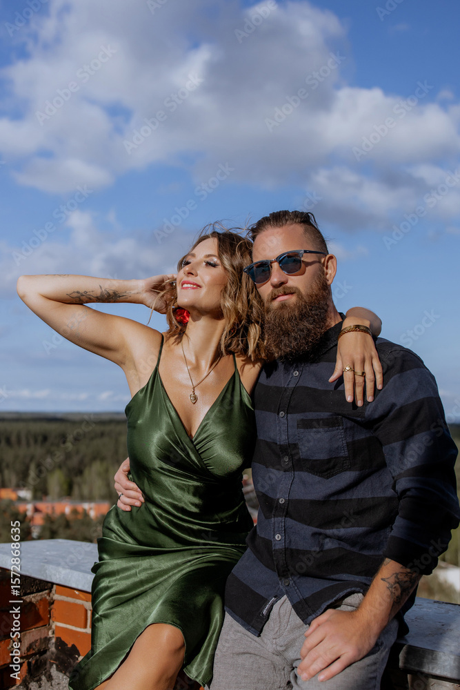 Naklejka premium Stylish Romantic Couple Sitting on Rooftop Embracing Each Other with Cityscape and Forest in Background — Urban Love and Fashion Concept