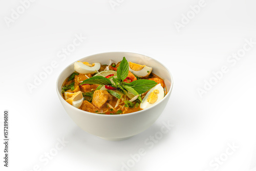 Delicious spicy Asian noodle soup with tofu, boiled egg, chili, lime, and herbs. Served in a white bowl on isolated background.