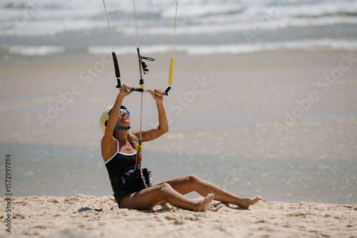 Woman learning kitesurfing on sandy beach, sitting with control bar during beginner training. Wearing helmet and sunglasses. First lesson.