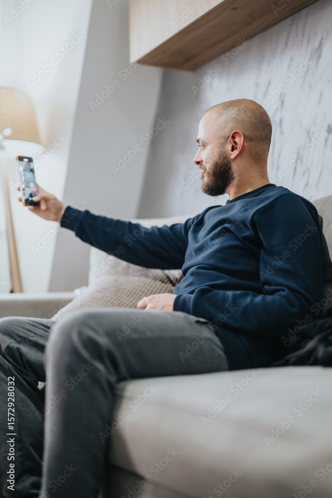Fototapeta premium A man with a beard sitting on a comfortable sofa indoors, capturing a selfie with his smart phone, enjoying a casual and relaxed moment near home decor.