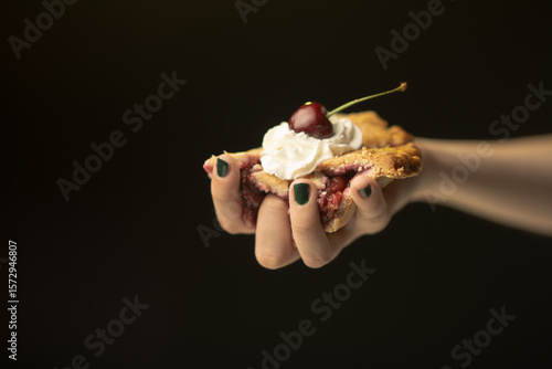 A close-up shot of a hand holding a slice of pie adorned with whipped cream and a cherry, illustrating the delicious textures and the joy of savoring culinary delights.