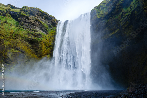 Skogafoss Waterfall displays a breathtaking plunge surrounded by verdant cliffs. Mist drifts in the air as sunlight filters through, creating a magical atmosphere in Icelands natural landscape.