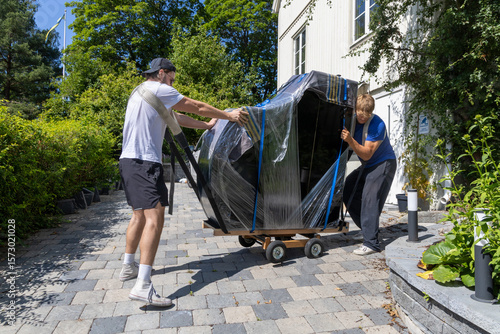 Two movers handling a wrapped grand piano on a dolly, navigating a paved path outside a residential building on a sunny day.