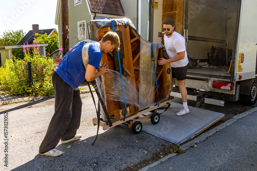 Two movers loading a partially wrapped grand piano onto a truck using a hydraulic lift on a residential street.