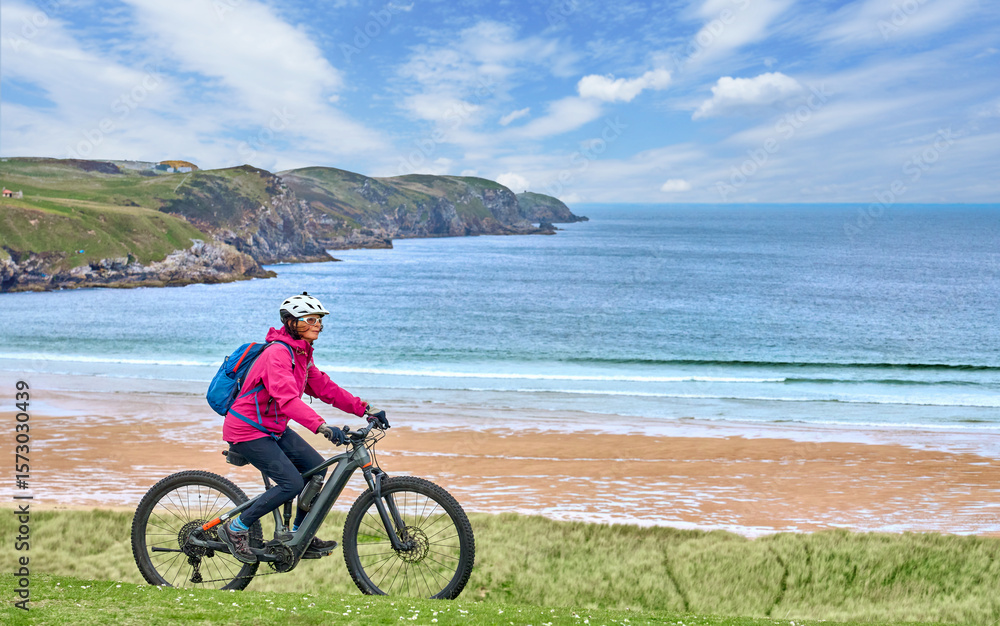 Fototapeta premium active senior woman riding her electric mountain bike on the cliffs of Strathy point lighthouse in Northern Scotland, UK 