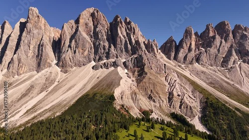 Breathtaking drone shot of dramatic Dolomite peaks rising above lush alpine forests and green valleys. Sunlight grazes the rocky summits of northern Italy's iconic UNESCO mountains