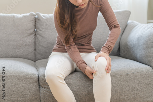 Schilderij op canvas Health care of knee pain concept, suffering asian young woman arthritis sitting joint ache, sore cramp or sprain tendon in leg on sofa at home