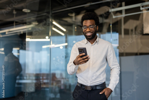 A smiling man in glasses looks at his phone in a modern office setting, appearing pleased with his device.
