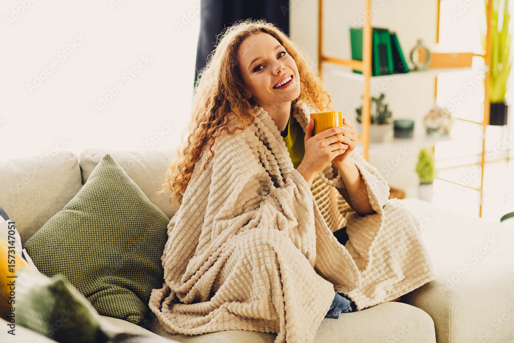 Fototapeta premium Young woman with curly hair enjoying a cozy moment at home, wrapped in a blanket, holding a mug and smiling.