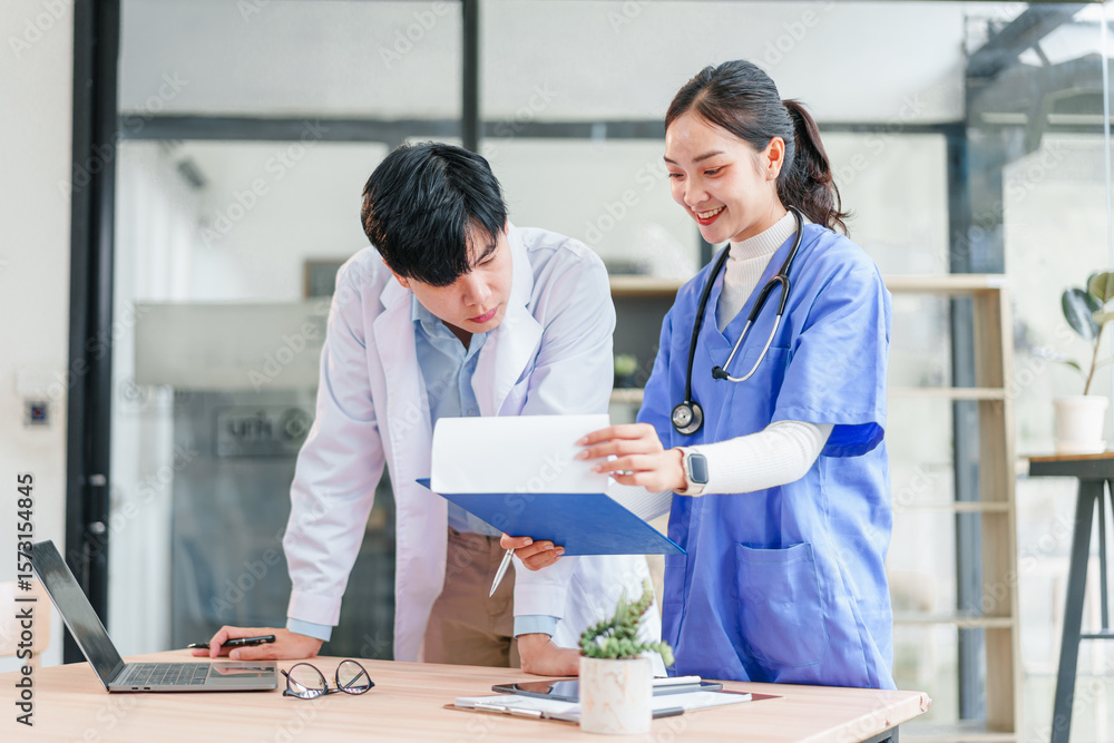 Fototapeta premium Two healthcare professionals, one male and one female, discuss patient records in bright modern office, both wearing medical uniforms and appearing focused and collaborative
