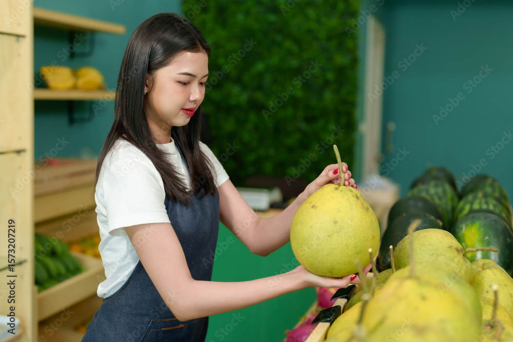 Fototapeta premium Asian female vendor lifting pomelo in hand checking quality of fruit inside fresh produce shop part of daily preparation routine of small tropical grocery business local vendor setup