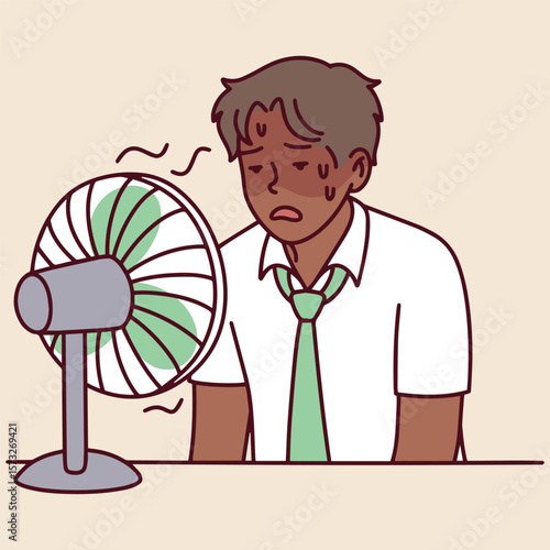 Dark-skinned man leans on table near small fan, visibly tired and sweating. He suffers from summer heat in indoor space