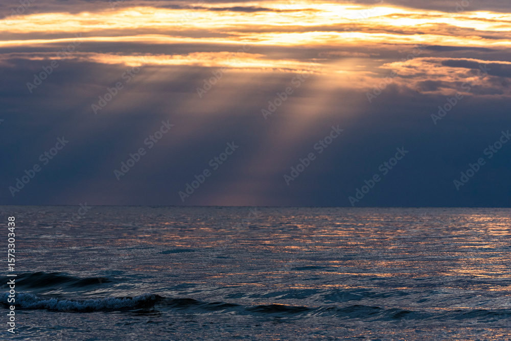 Obraz premium Calm sea under a cloudy sky at sunset. Sunlight filters through the clouds, creating rays over the water. 