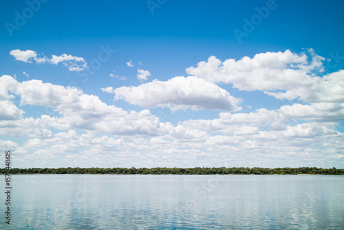 Natural landscape with a calm river in the foreground, reflecting the blue sky. On the horizon, there is a line of leafy green trees. It is filled with evenly dispersed, fluffy white clouds