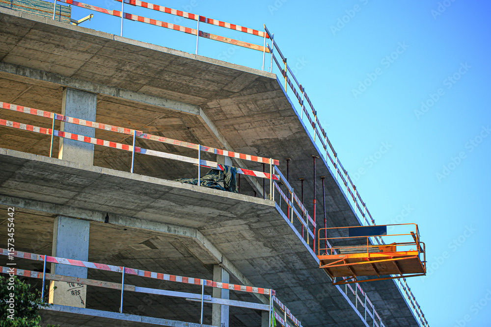 Naklejka premium Modern concrete building under construction with orange suspended lift platform and red safety railings. Clear blue sky background highlights the architectural geometry and urban development