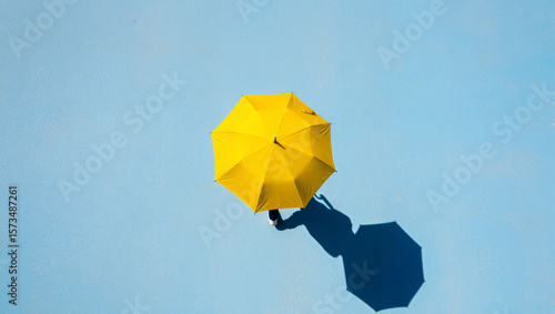 A person walks under a bright yellow umbrella casting a long shadow on a clear blue surface.