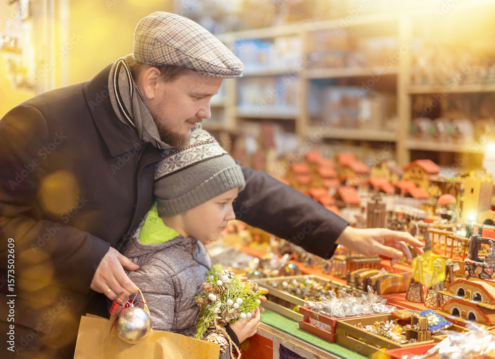 Fototapeta premium At fair with festive goods, man and son near counter with decorations and figures for nativity scenes, buy ceramic products, toy carts and figures of peasants. .