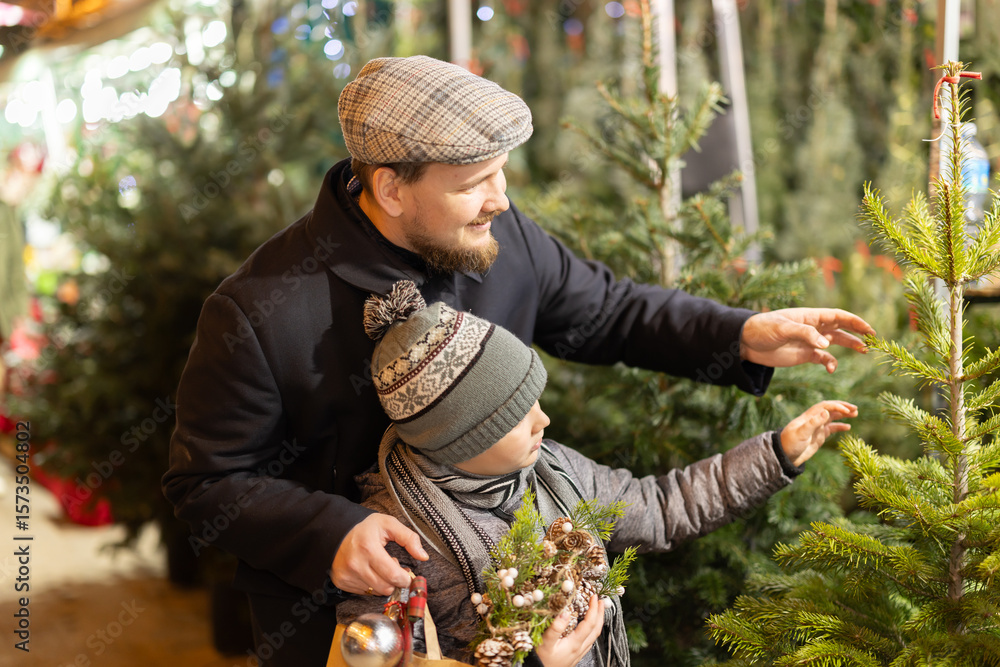 Fototapeta premium Dad and son are looking at natural and artificial Christmas trees at street fair. Family walks around bazaar, examines goods, and stares at festive assortment of fair. .