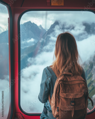 Woman with Backpack Looking Out Gondola Window. Young woman with a backpack gazes out from a red gondola cabin, admiring the misty mountain peaks and clouds below.

