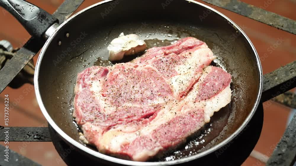 A close-up shot of a raw, seasoned ribeye steak being pan-seared. Garlic cloves are added, followed by a generous pat of butter, illustrating the process of cooking a delicious, juicy steak. A close-up shot of a raw, seasoned ribeye steak being pan-seared. Garlic cloves are added, followed by a generous pat of butter, illustrating the process of cooking a delicious, juicy steak.