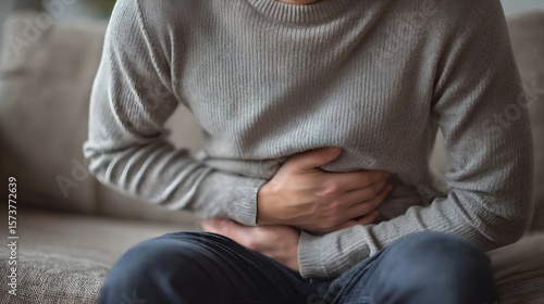 Fototapeta Naklejka Na Ścianę i Meble -  Close up view of a person s hands on their sto h indicating a gastro infection indigestion or constipation while resting on a sofa at home